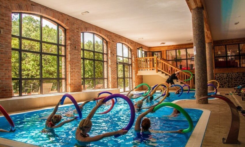 Indoor hotel pool with arched brick windows, group aqua fitness class using colorful pool noodles led by instructor.