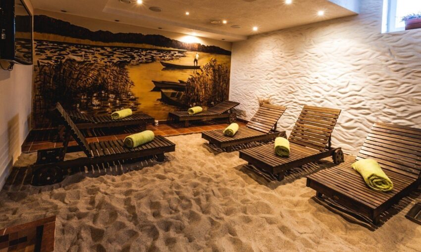 Indoor spa relaxation room with sand floor, wooden lounge chairs each with rolled green towel, warm mural of boats and reeds.