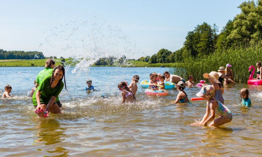 Lakeside family beach with children and adults splashing in shallow water, colorful inflatables, reeds and a sunny countryside view