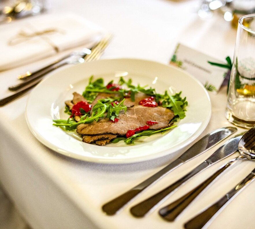 Thinly sliced roast beef on arugula with red berry sauce, elegant white-tablecloth place setting and glass