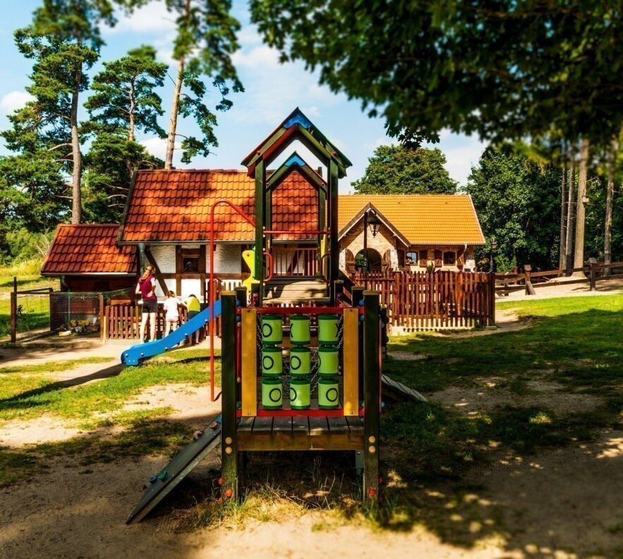 Plac zabaw z zjeżdżalnią i drewnianą wieżą, dzieci bawią się przy niej, przed rustykalnymi domkami z czerwonymi dachami i sosnami