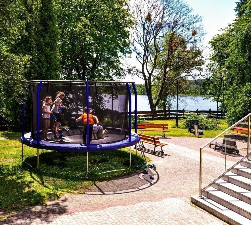Ogród hotelowy nad jeziorem z dziećmi skaczącymi na ogrodzonej trampolinie, drewnianymi ławkami i widokiem na wodę.