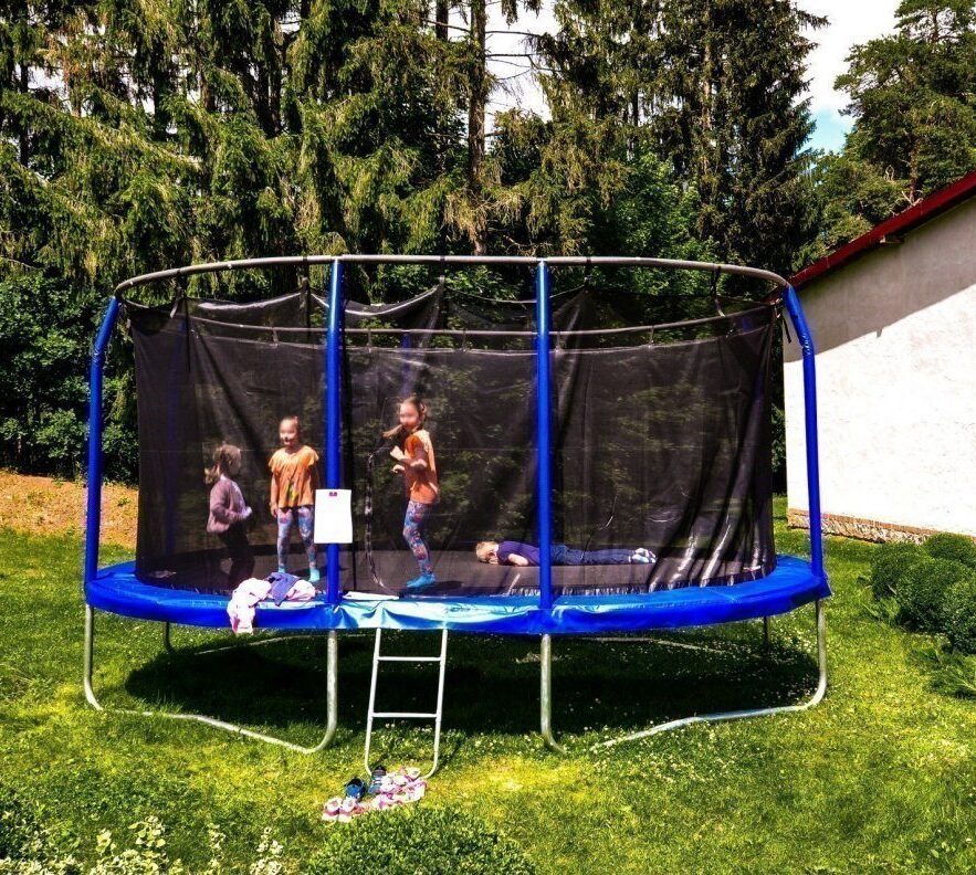 Trampolina z siatką na trawniku, dzieci skaczą i bawią się; drabinka i buty przy wejściu