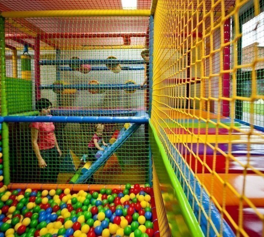 Colorful indoor kids' playroom with ball pit, padded multi-level climbing frames, slide and an adult supervising a child.