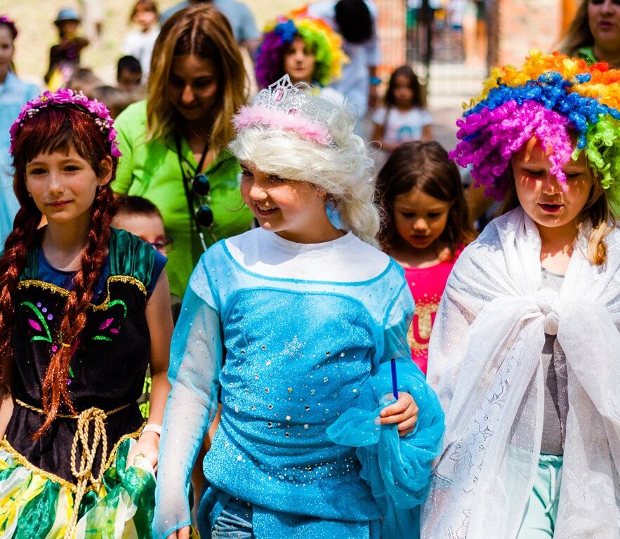 Group of children in colorful costumes and wigs walking outdoors during a lively family-friendly costume parade