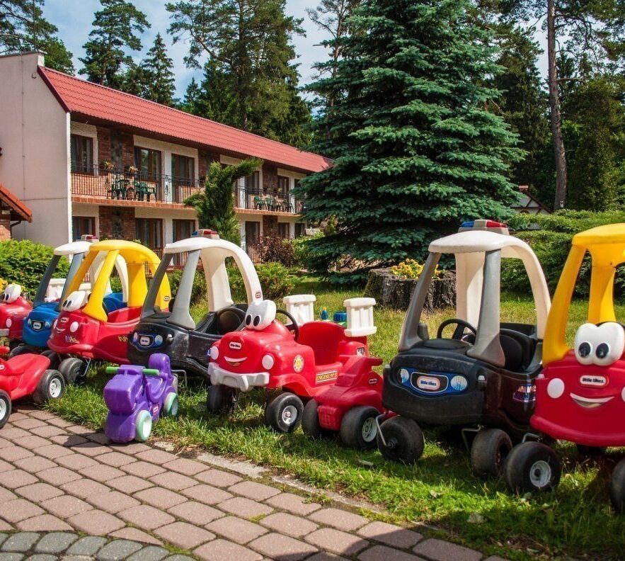 Rząd kolorowych dziecięcych samochodzików na trawniku przed rodzinnym hotelem z balkonami i sosnowym ogrodem