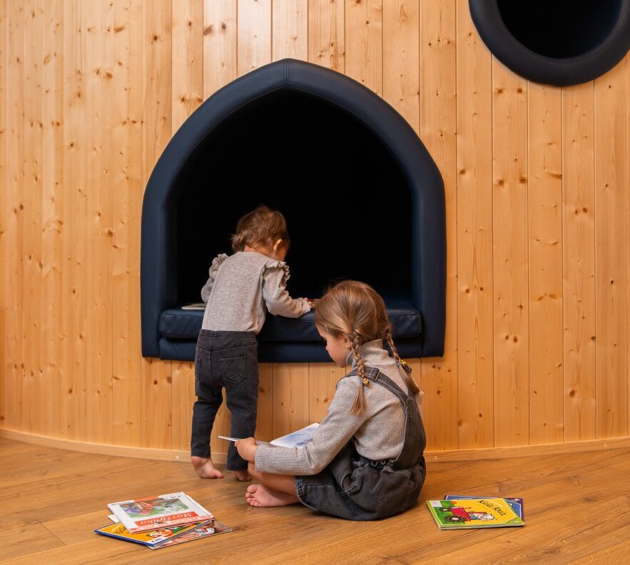 Wood-paneled children's play corner with a padded navy reading nook; two children reading books on the floor.
