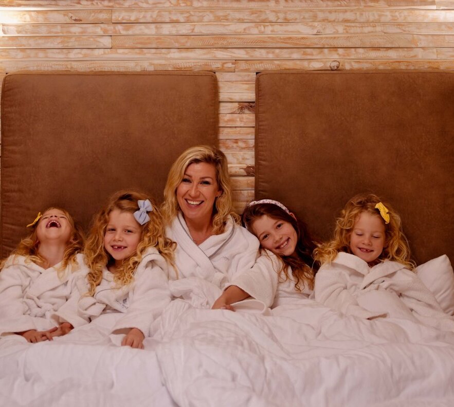 Family relaxing in a plush hotel bed wearing white robes, smiling in a cozy room with a wood-accented wall