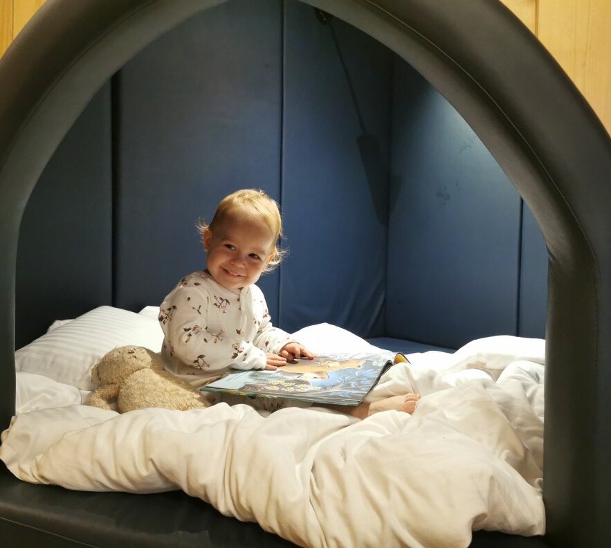 Toddler smiling in a cozy canopy bed nook with plush bedding, storybook and teddy - family-friendly hotel room