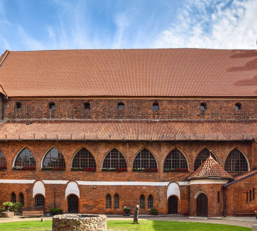 Historic red-brick courtyard with arched windows and flower boxes, red-tile roof, stone well, benches and green lawn.
