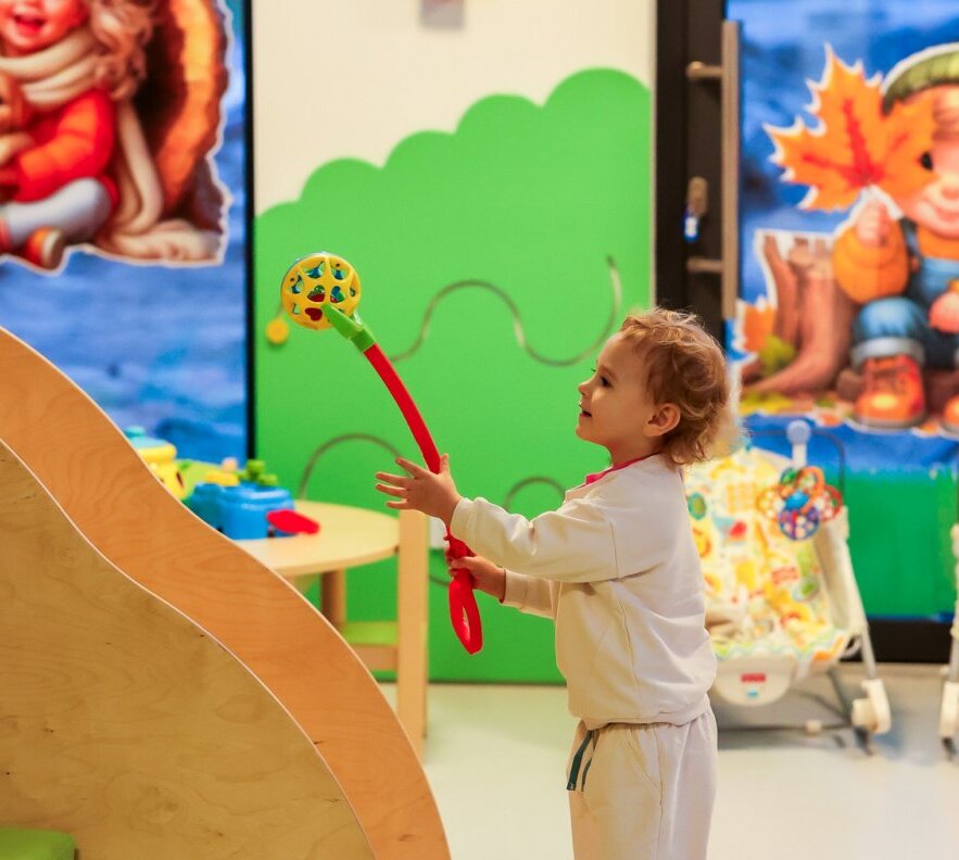 Toddler playing with a colorful rattle in a bright hotel kids' playroom with murals, activity table and baby bouncers.
