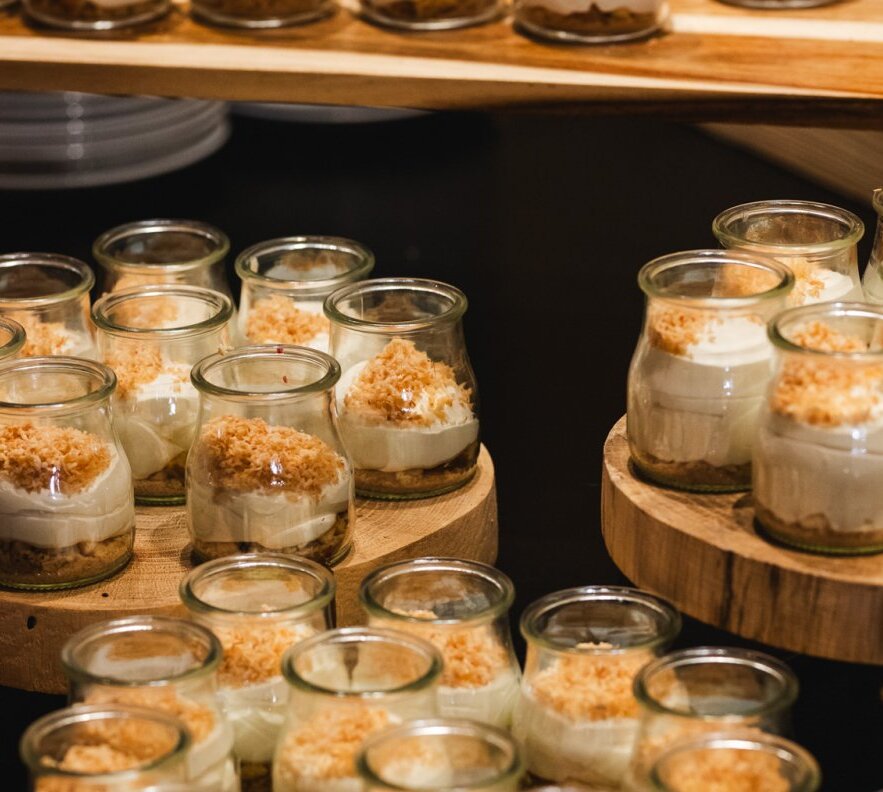 Individual layered cream desserts in small glass jars topped with golden crumble, displayed on wooden risers at a hotel buffet.