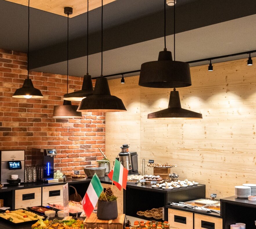 Buffet with pastries, cold cuts and coffee station beneath industrial pendant lights, exposed brick and wood walls, small Italian flags.