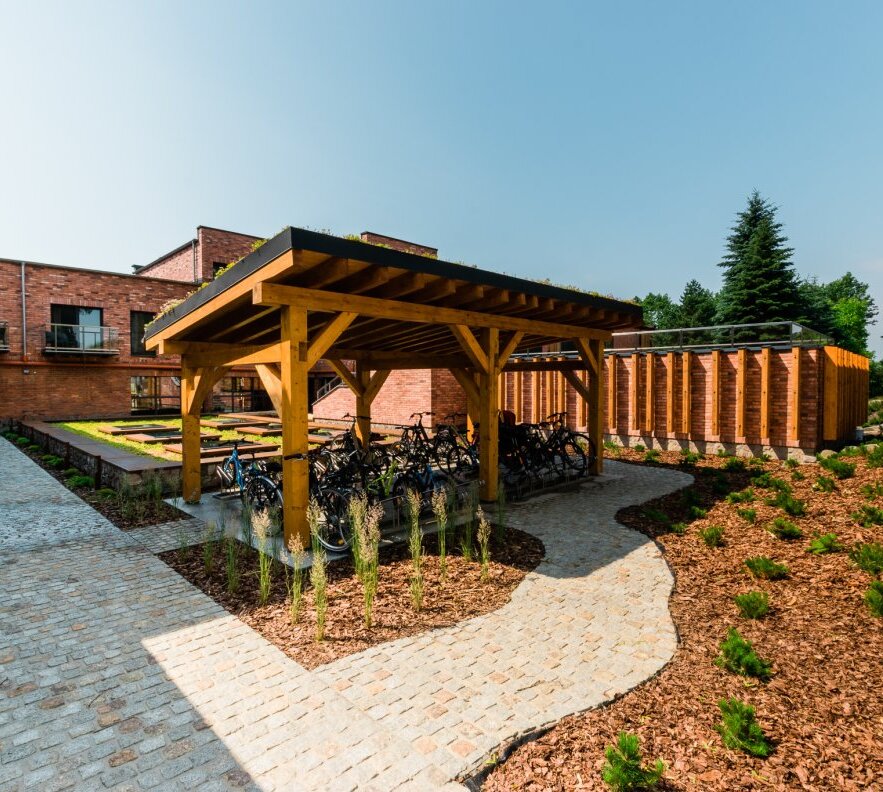 Covered wooden bike shelter with multiple bicycles, paved courtyard, brick hotel buildings and landscaped garden beds