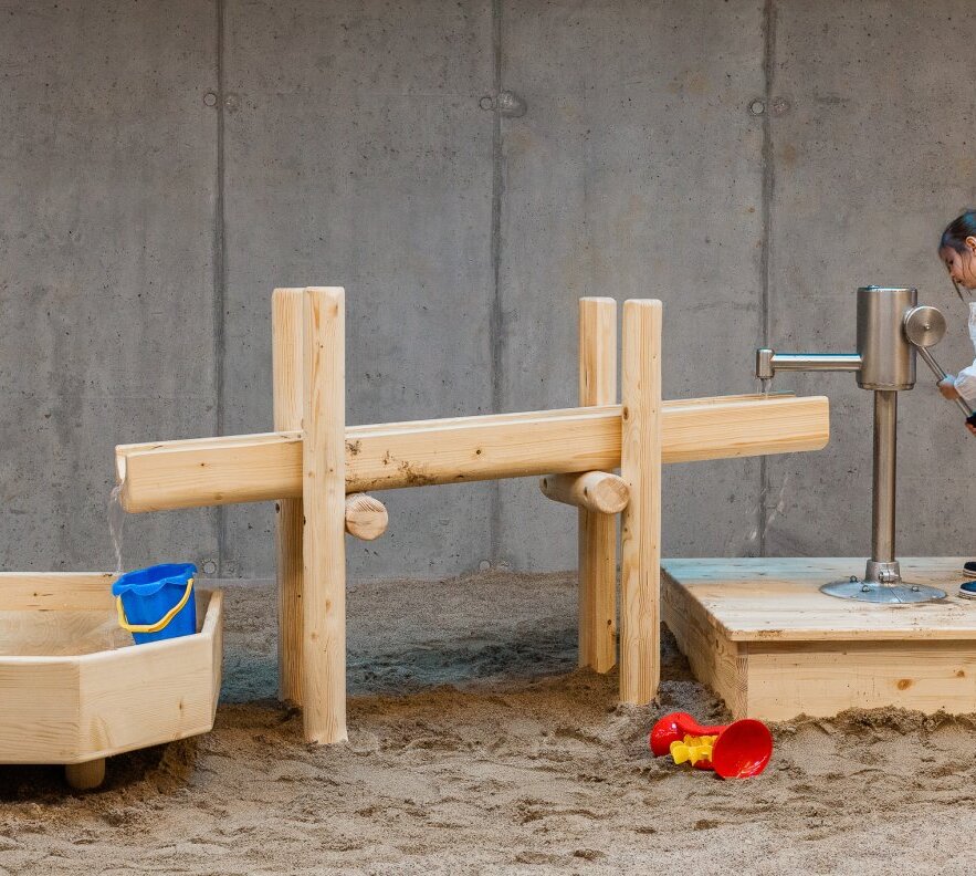 Indoor children's sand play area with wooden water channels and hand pump; child pumping water, sandpit with blue bucket and red shovel.