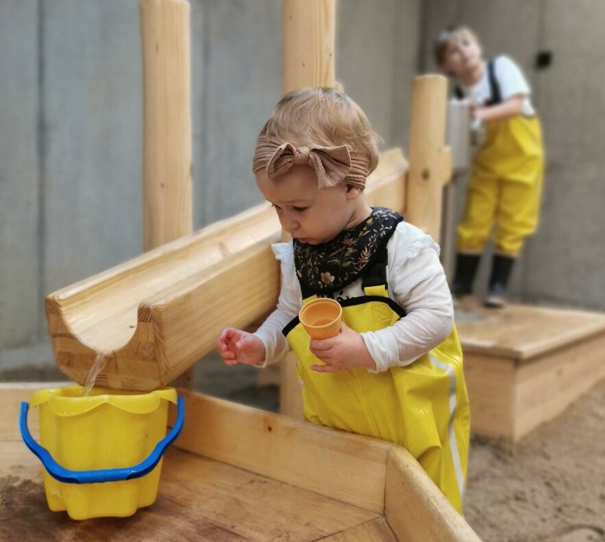 Toddler in yellow waterproof overalls and bow headband scoops water with a cup into a yellow bucket from a wooden water play table; another child behind.