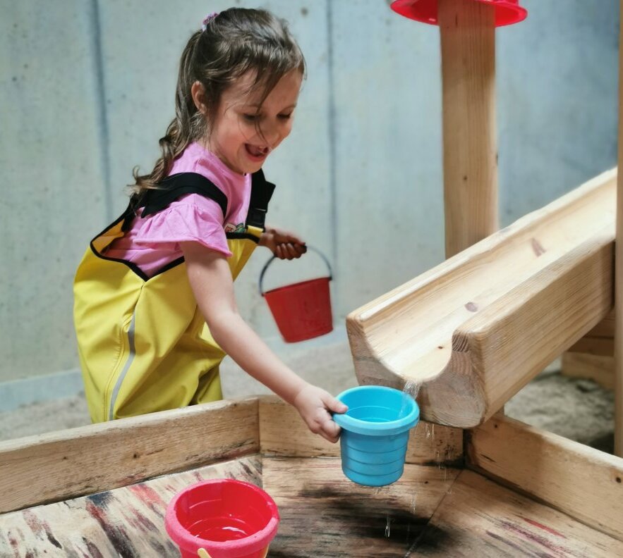 Child in yellow waterproof overalls filling a blue bucket from a wooden water channel in an indoor children's play area.