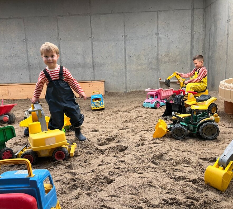 Indoor sand play area with concrete walls and wooden edge; two children in waterproof overalls playing with toy excavators and trucks