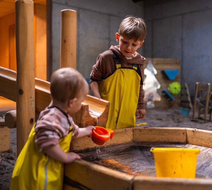Two children in yellow waterproof overalls playing with water and sand at a wooden play table in a family-friendly indoor playroom