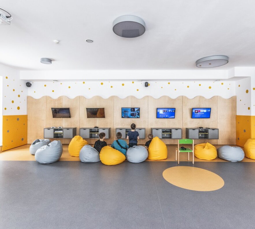 Family game room with wall-mounted TVs and consoles, children playing on yellow and gray beanbags in bright, modern space.