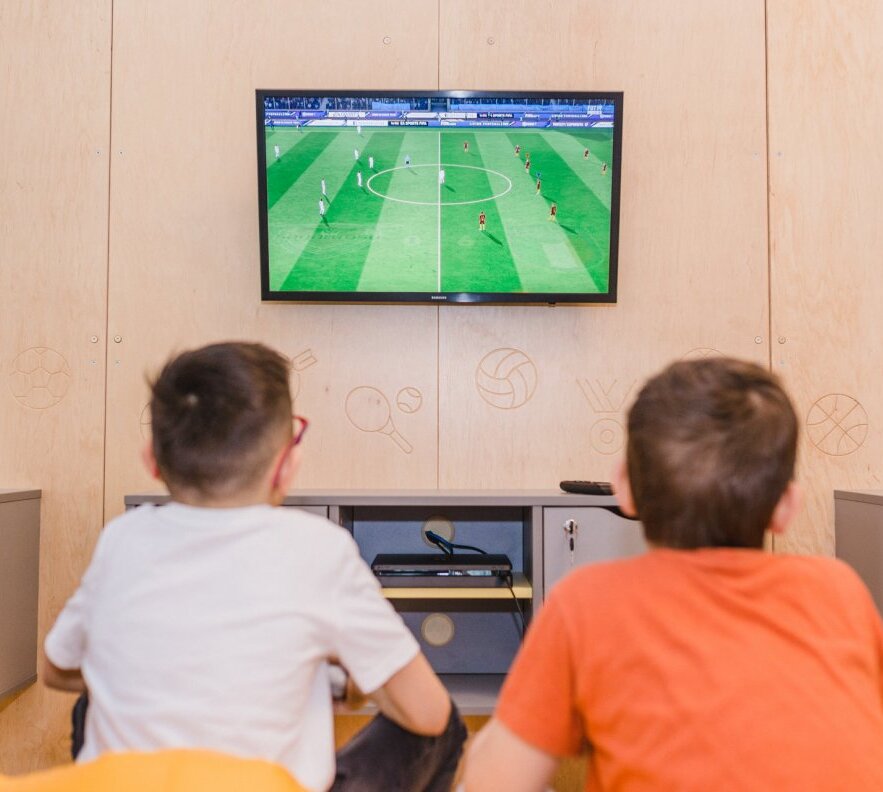 Two boys on beanbags watching a soccer match on a wall-mounted TV in a family game room with wood-panel sports icons and console