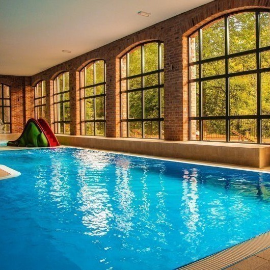 Indoor heated pool with arched brick windows, natural light, bench seating and a small children's slide overlooking trees.