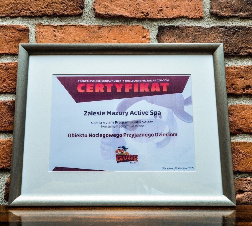 Oprawiony certyfikat na ceglanej ścianie potwierdzający, że Zalesie Mazury Active Spa to obiekt przyjazny dzieciom