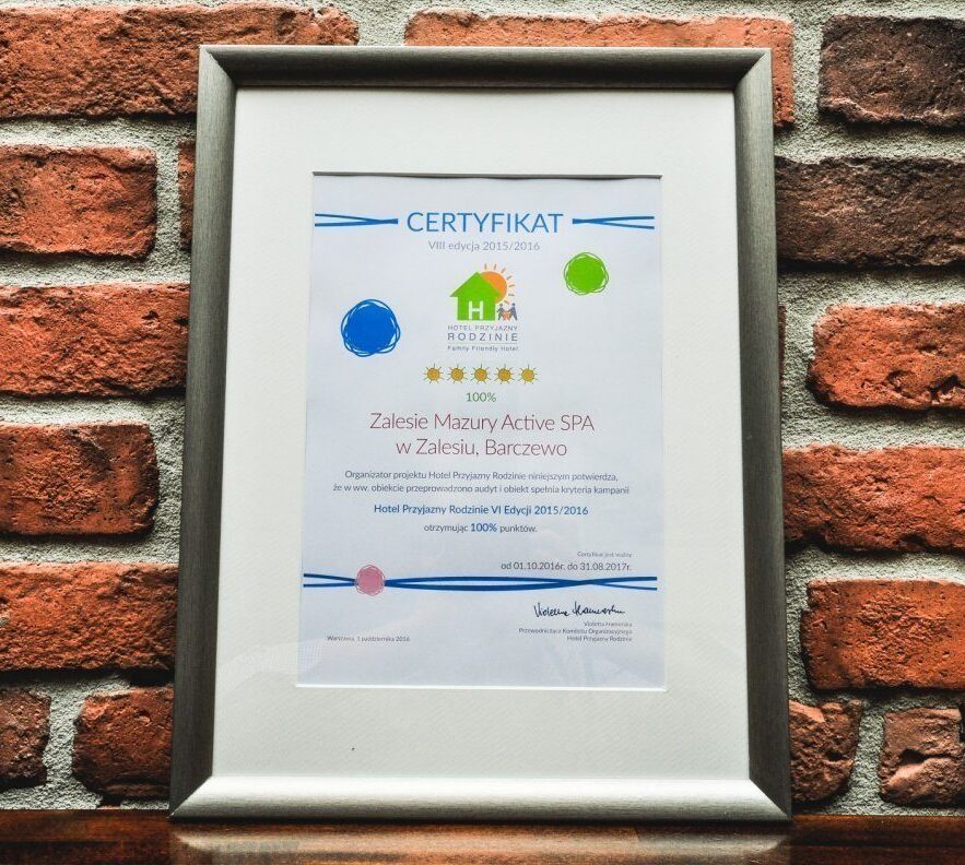 Oprawiony certyfikat Zalesie Mazury Active SPA na surowej ceglanej ścianie, z ikonami pięciu gwiazdek i oznaczeniem 100%.
