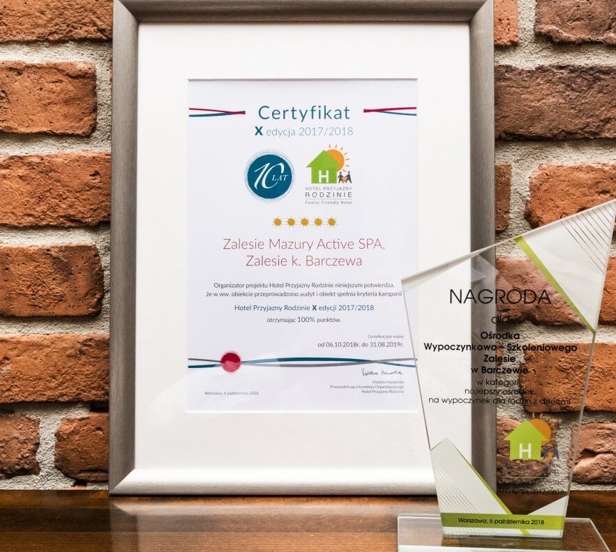 Oprawiony certyfikat i szklana statuetka na tle ceglanej ściany, nagradzające Zalesie Mazury Active SPA jako rodzinne spa.