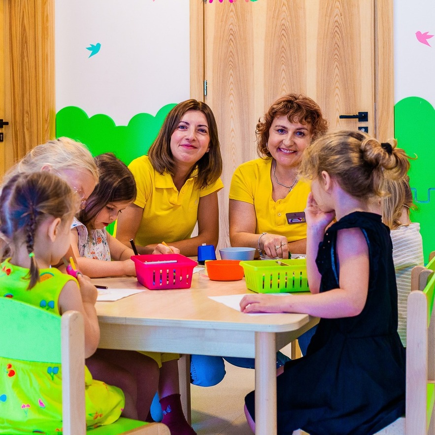 Hotel kids' playroom craft session - two staff in yellow supervising children at a wooden table with colorful bins