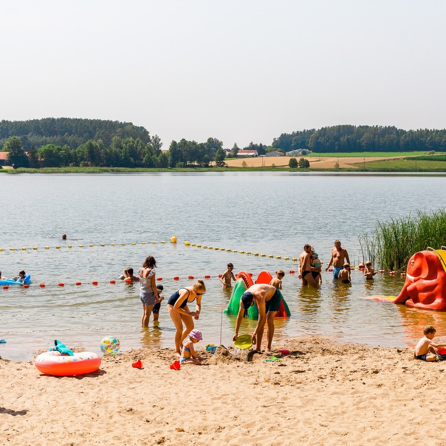 Family lakeside beach with sandy shore, buoyed shallow swim area, kids playing and a small red water slide.