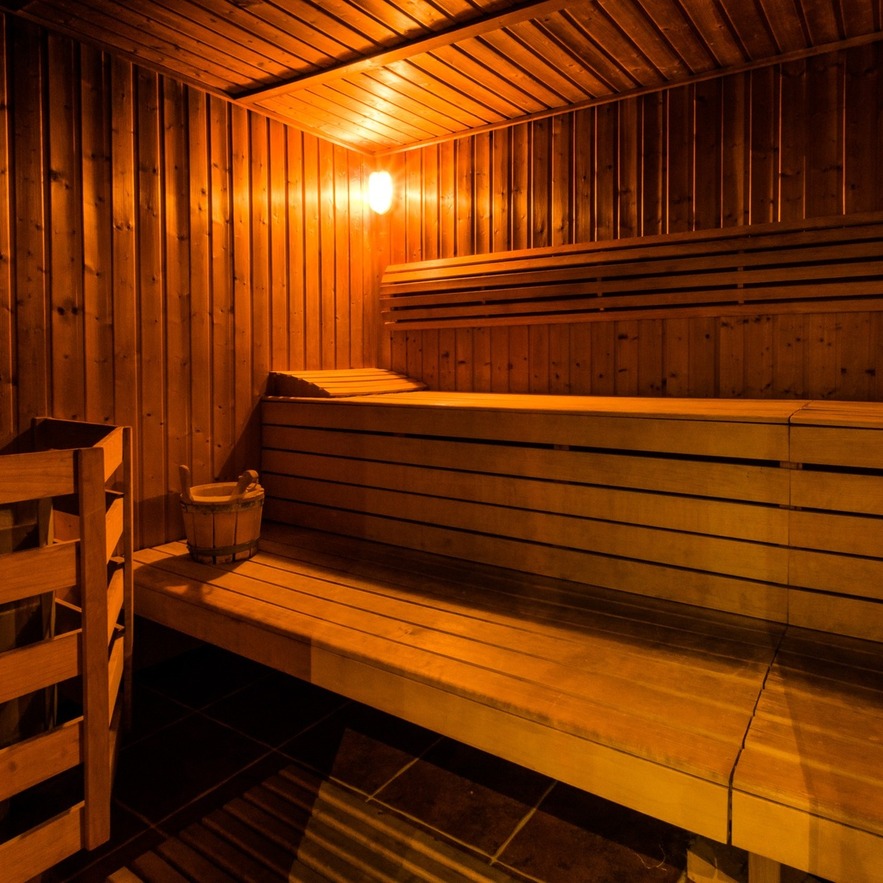 Sauna hotelowa obita drewnem, piętrowe ławy, piec z kamieniami i wiadro, łagodne bursztynowe światło, relaks w spa.