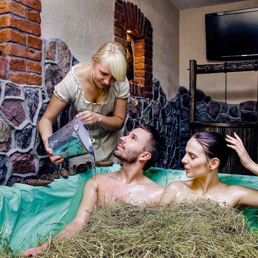 Couple enjoying a traditional hay bath in a rustic spa tub; attendant pours water beside stone-and-brick walls