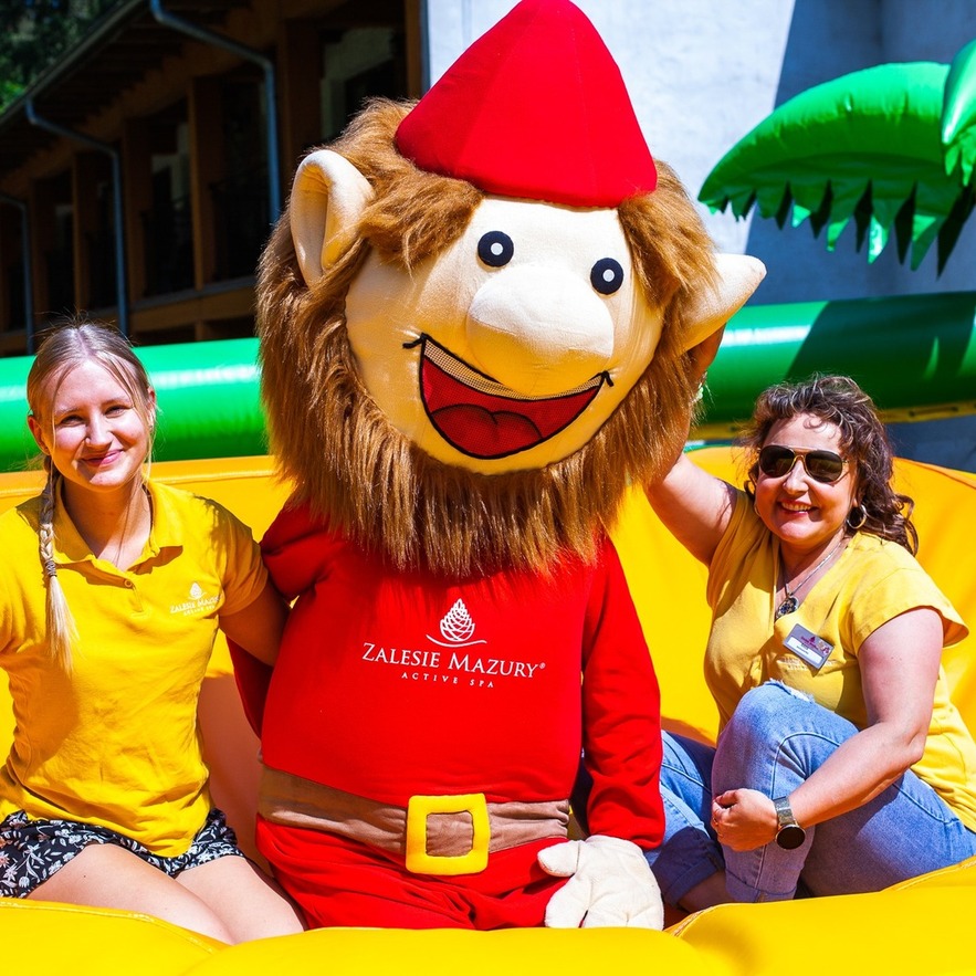 Resort staff smile beside a red-costumed mascot on a bright yellow inflatable play area — family-friendly kids' entertainment at Zalesie Mazury