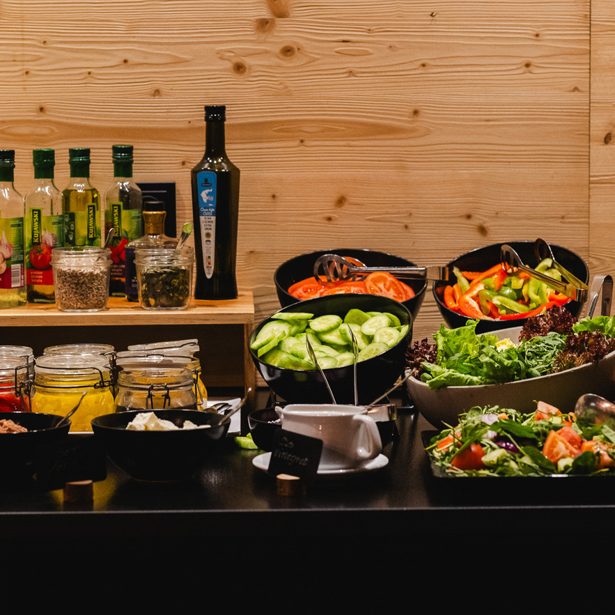 Self-serve salad buffet with bowls of cucumber, tomatoes, mixed greens, dressings, oils and jars on a wooden counter