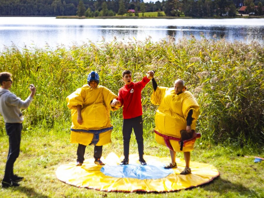 Team building na zewnątrz: goście w dmuchanych strojach sumo na macie nad jeziorem, prowadzący podnosi rękę zwycięzcy, inny fotografuje
