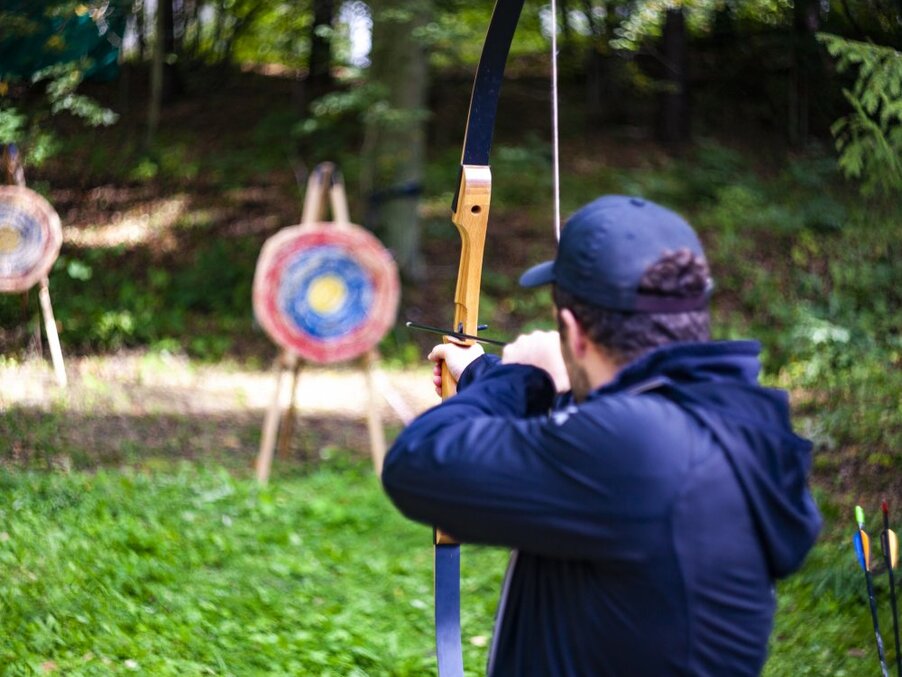 Guest aiming a recurve bow at straw targets on a grassy, wooded outdoor archery range