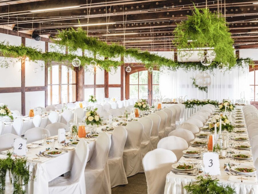 Elegant rustic reception with long white tables, chair covers, floral centerpieces and suspended greenery under wooden beams