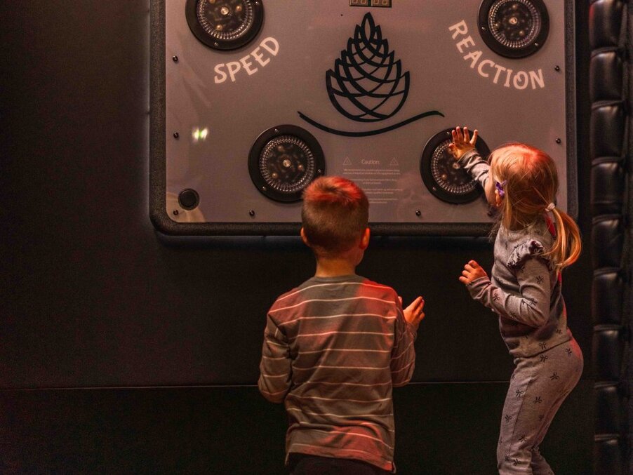 Two children play a wall-mounted "Speed/Reaction" game in a dim family entertainment area; a girl reaches to touch a lit sensor.