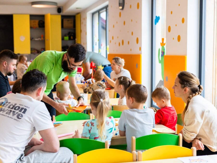 Jasny klub dziecięcy w hotelu z dużymi oknami, kolorowymi krzesłami, stołami do zajęć i animatorem pomagającym dzieciom.