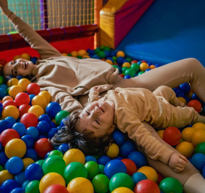 Family play area with colorful ball pit; parent and child relaxing and laughing on padded play structure