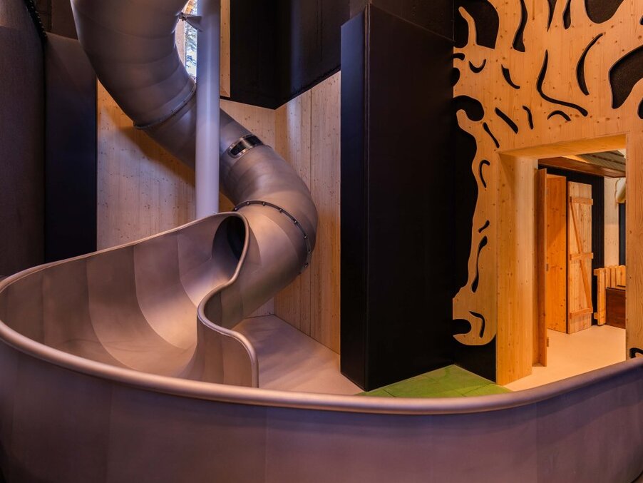 Indoor spiral metal slide winding through a warm wood-clad family play area with carved wooden panels and soft mood lighting