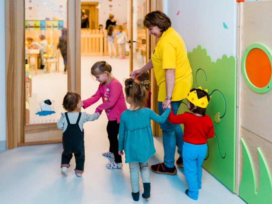 Hotel kids' club: caregiver in yellow holding hands with toddlers entering a bright, colorful playroom with toys and activity tables