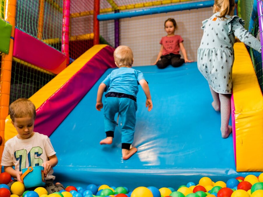Family-friendly indoor play area with bright slide and ball pit; children climbing, sliding and playing.