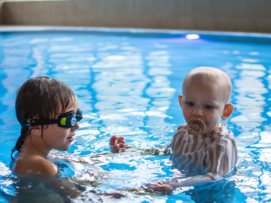 Indoor hotel pool with children: older child in goggles teaching younger baby with pacifier to enjoy shallow family-friendly swimming area.