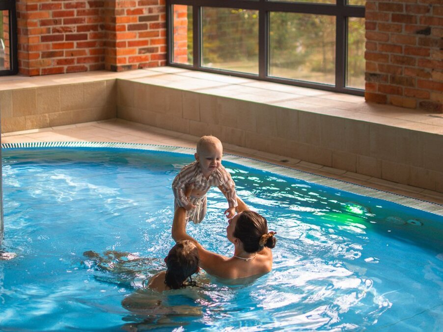 Woman lifts a baby above the water in a bright indoor hotel pool with exposed brick walls and large windows