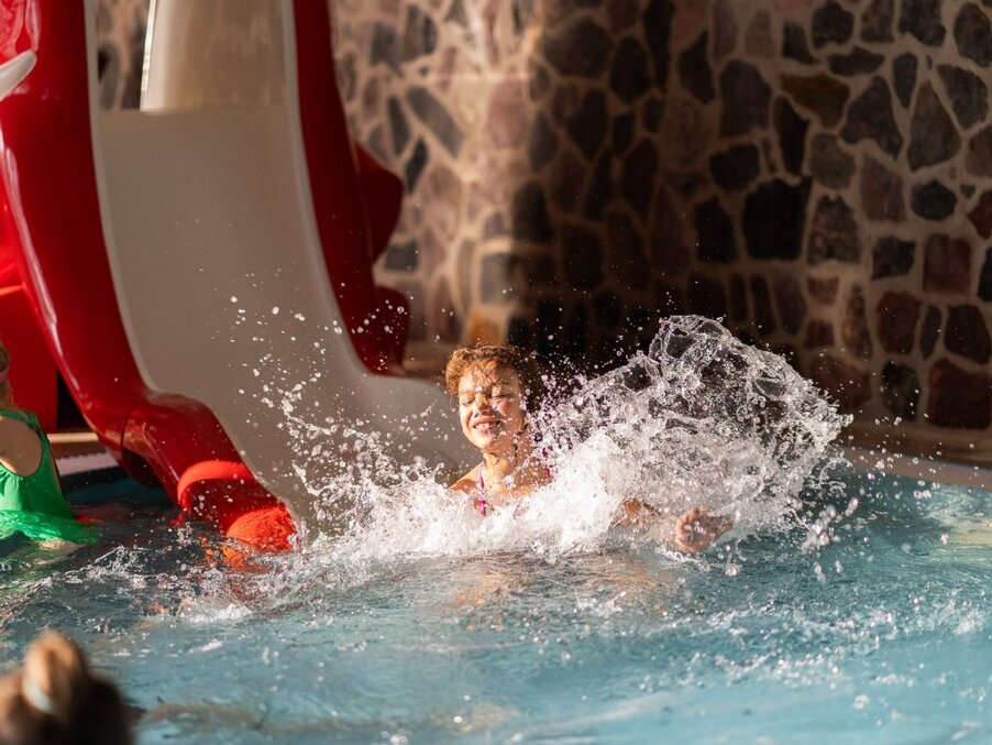Child splashing into an indoor pool from a red waterslide, sunlit family pool area with stone wall backdrop