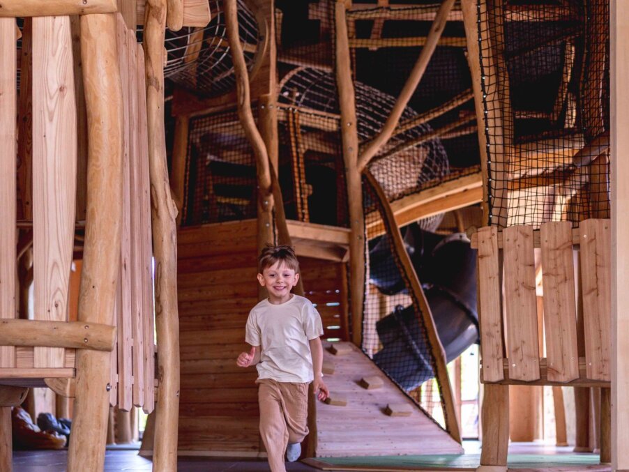 Wooden indoor playground with netted slides and climbing ramp; smiling child running along a hotel play structure