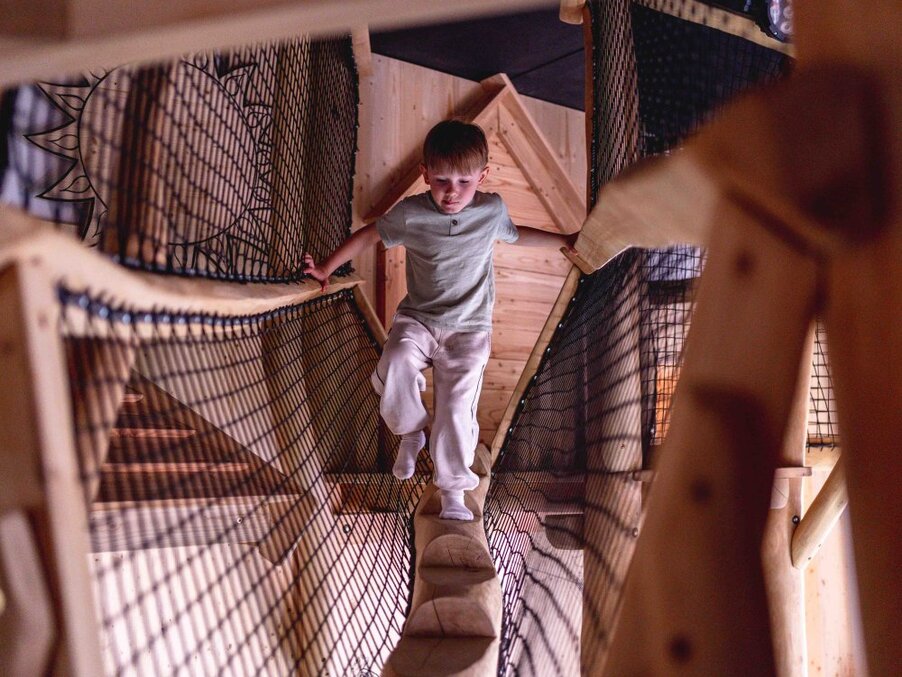 Child walking along a wooden balance beam inside an indoor play structure with safety netting and warm timber walls