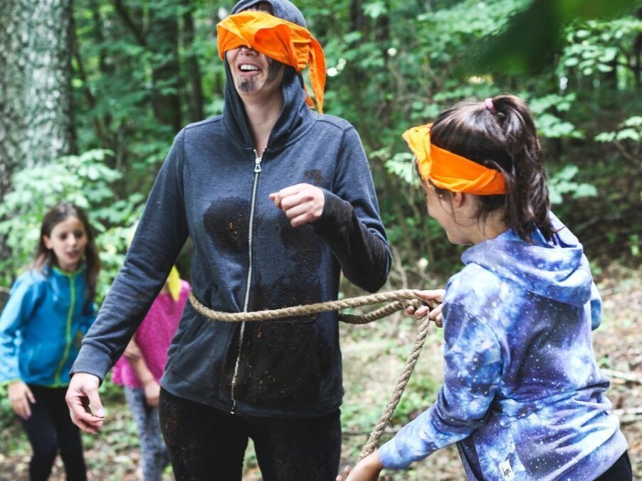 Blindfolded adult with an orange band guided by children holding a rope during a playful forest team-building game.